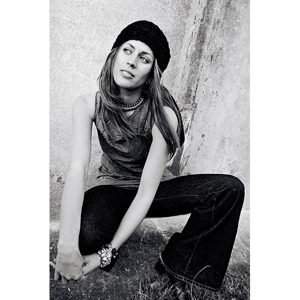A model squats in front of a concrete wall. She has long hair and wears a beanie a halter top and dark bootleg jeans.