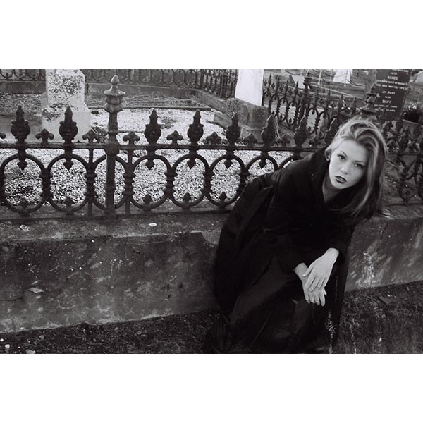 black and white photo in a cemetary. A model sits on a concrete wall and leans forward. She wears a black jacket. The wall is topped with a wrought iron fence. There are headstones behind the wall.